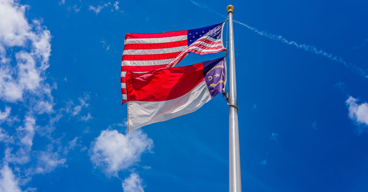 American and North Carolina state flags waving against a clear blue sky with scattered clouds.